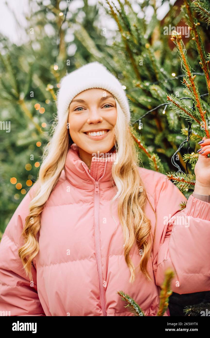 Une jolie blonde choisit un arbre de Noël pour la nouvelle année. Une femme dans un chapeau et une veste rose sourit à l'appareil photo par temps enneigé. Vacances d'hiver et n Banque D'Images