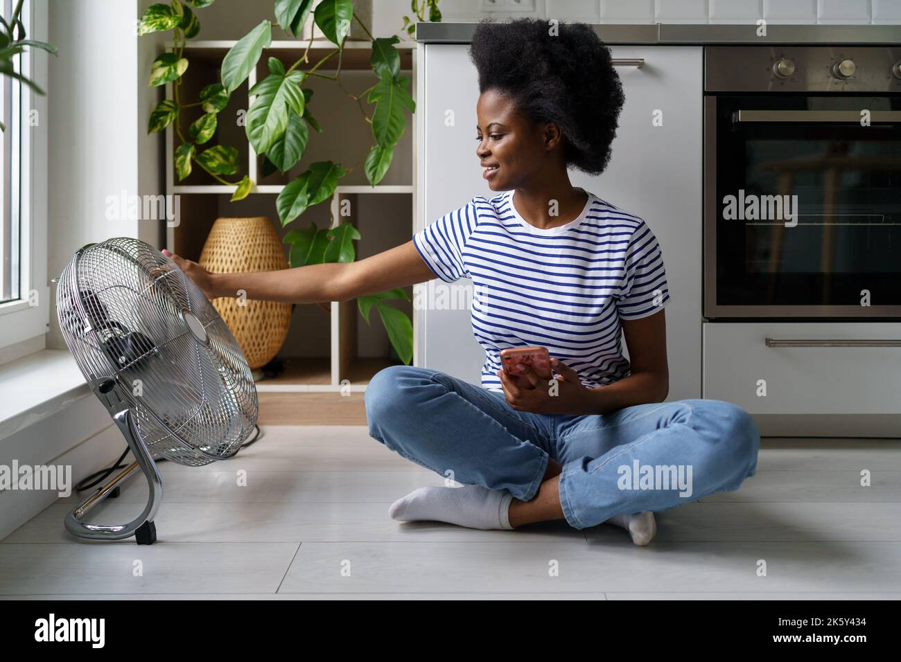 Femme afro-américaine satisfaite appréciant l'air frais provenant de derrière le ventilateur électrique et tient le téléphone Banque D'Images