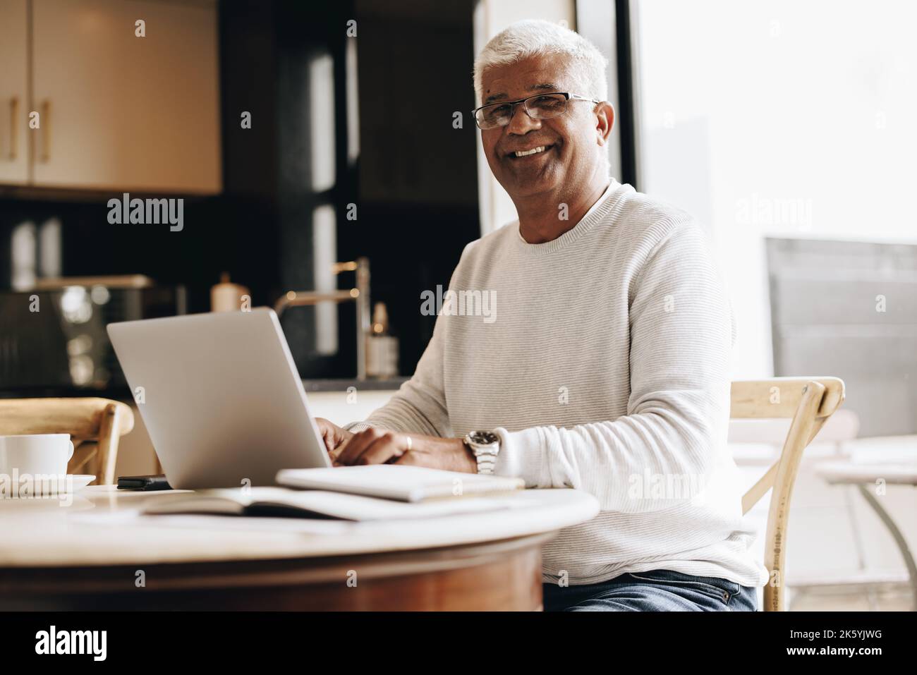 Un homme d'affaires expérimenté souriant à l'appareil photo tout en étant assis avec un ordinateur portable dans son bureau à domicile. Homme d'affaires mature saisissant un e-mail professionnel pendant son travail Banque D'Images