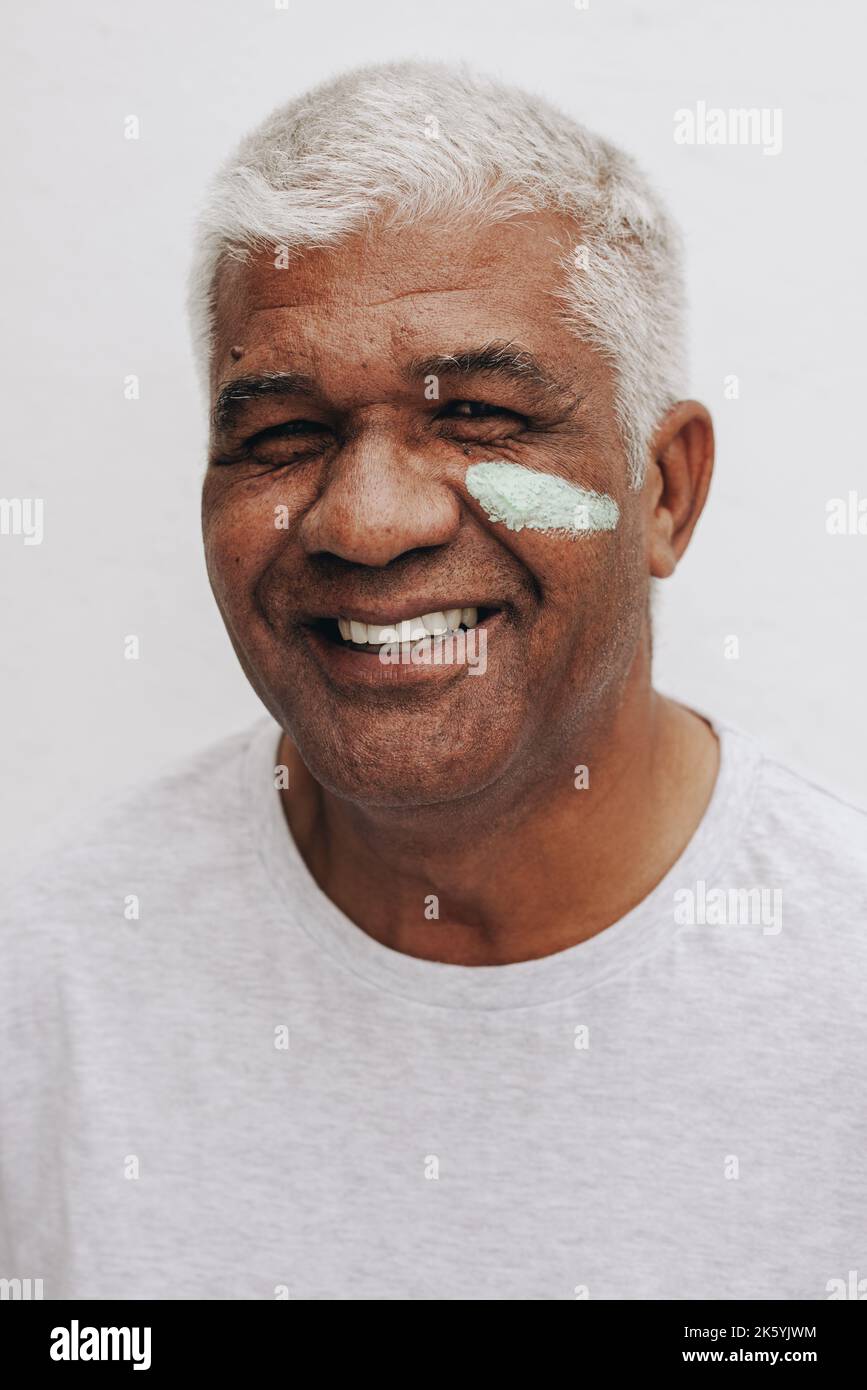 Joyeux homme mûr souriant à l'appareil photo avec de la crème hydratante sur son visage. Homme senior positif pour le corps traitant sa peau vieillissante avec une beauté nourrissante Banque D'Images