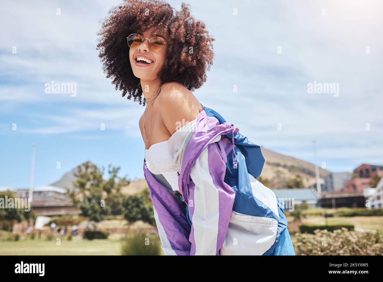 Portrait d'une jeune femme branchée de race mixte avec un afro souriant et posant seul à l'extérieur. Femme hispanique portant des lunettes de soleil et se sentant heureuse Banque D'Images