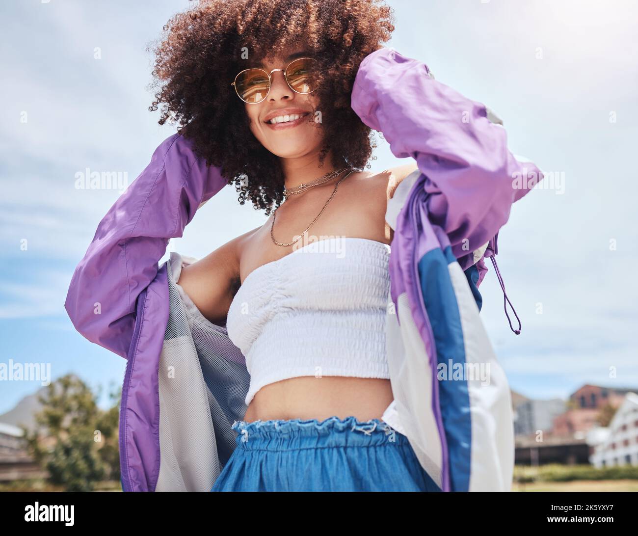 Portrait d'une jeune femme branchée de race mixte avec un afro souriant et posant seul à l'extérieur. Femme hispanique portant des lunettes de soleil et se sentant heureuse Banque D'Images
