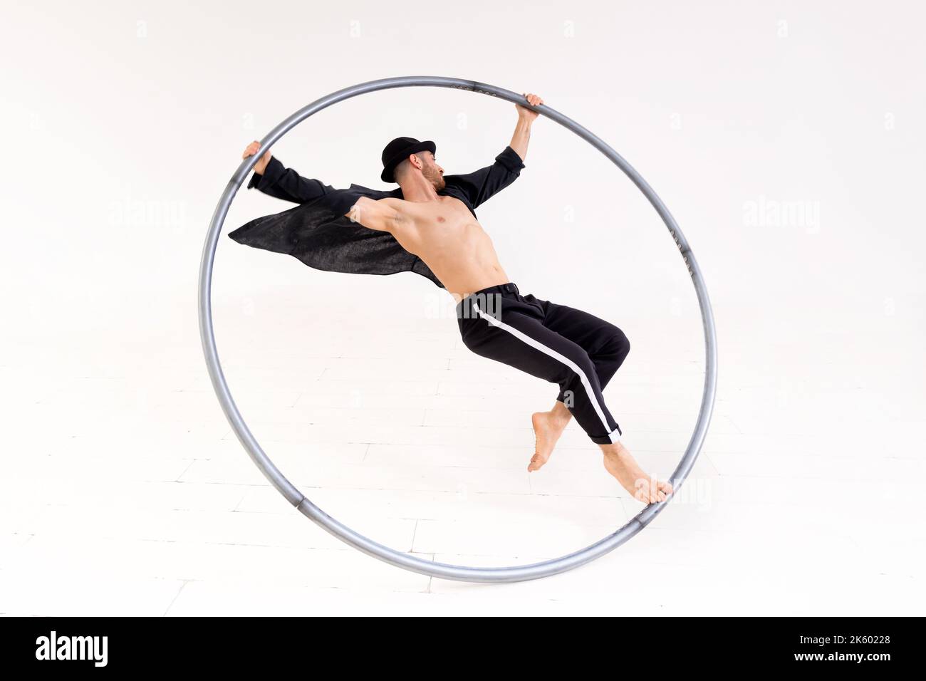 Tout le corps d'acrobat mâle pieds nus regardant loin et exécutant le stunt sur la roue Cyr pendant la répétition sur fond blanc Banque D'Images