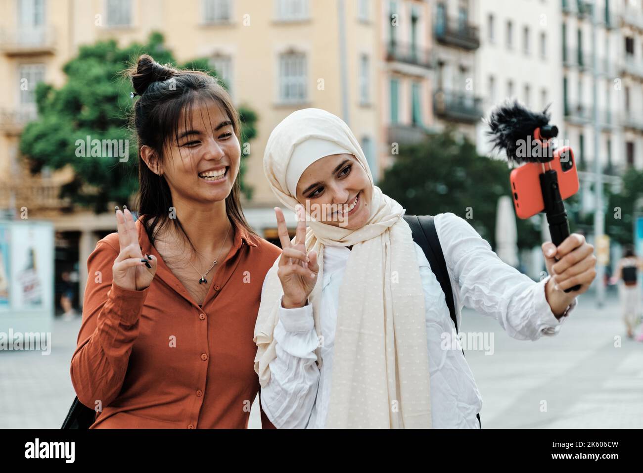 Amis multiethniques souriant et faisant signe de paix avec les doigts tout en streaming avec un téléphone mobile à l'extérieur. Banque D'Images