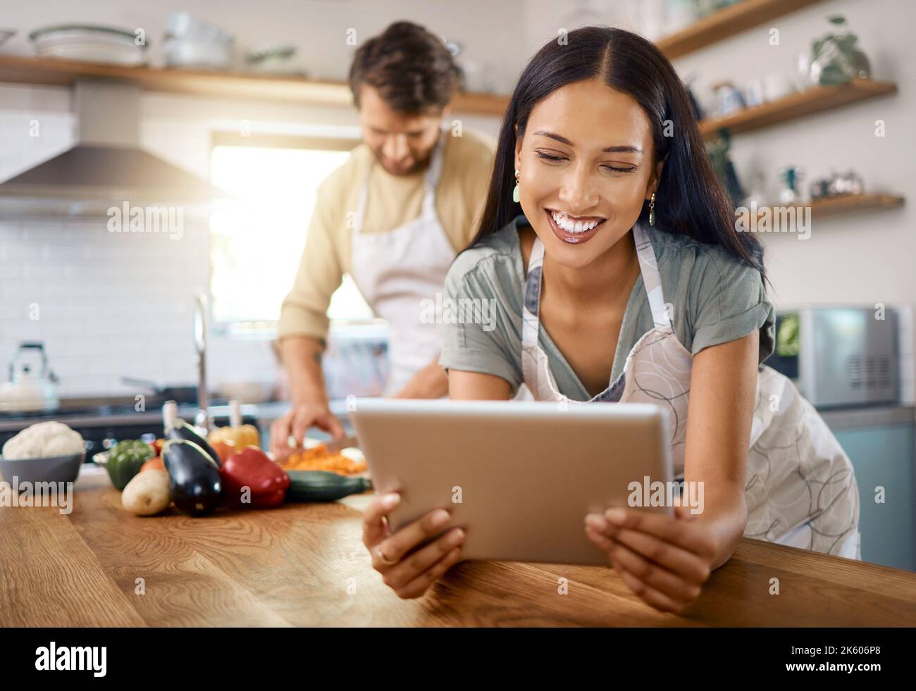 Gros plan sur une jeune femme heureuse qui navigue sur une tablette numérique tandis que son petit ami cuisine en arrière-plan. Jeune femme hispanique à la recherche de recette à Banque D'Images