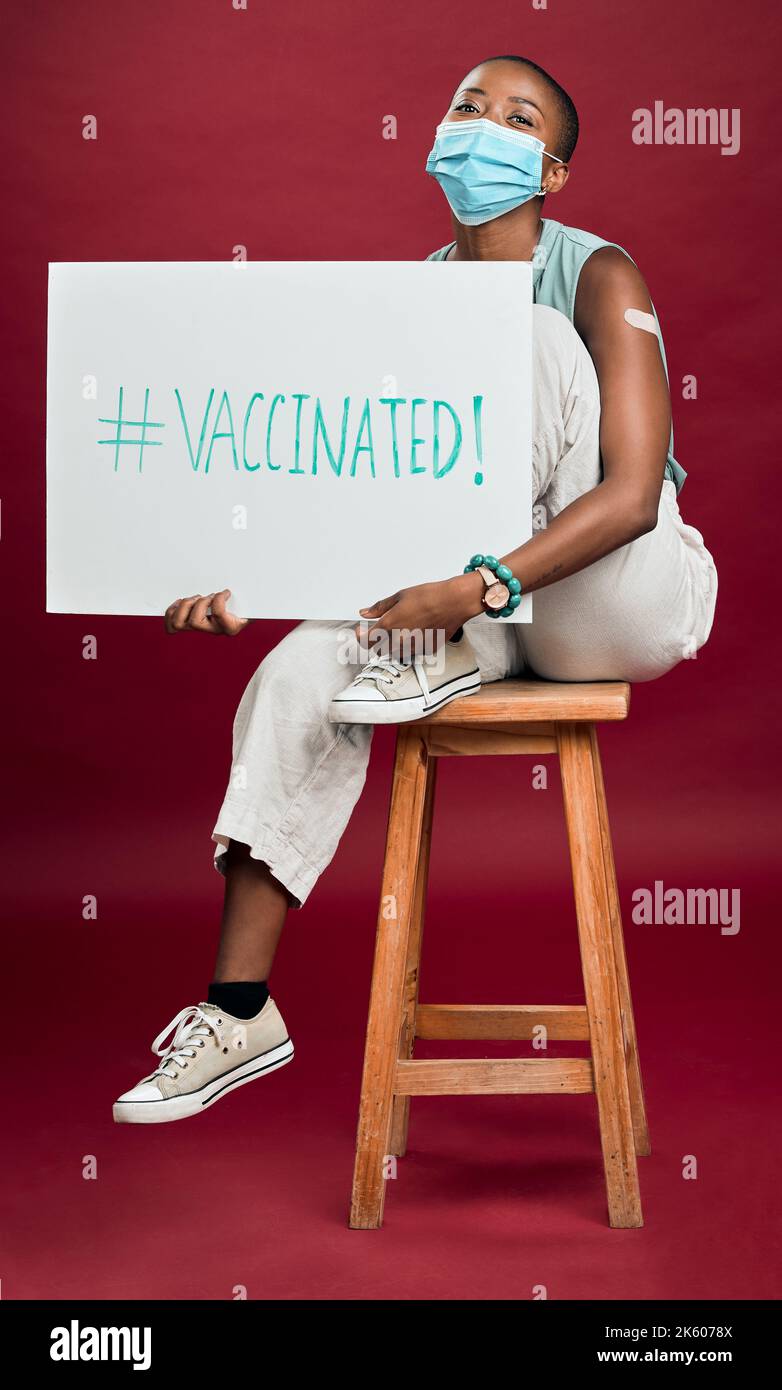 Femme vaccinée Covid afro-américaine montrant du pansement pour bras, tenant une affiche, portant un masque chirurgical. Happy Black model isolé sur Red studio Banque D'Images