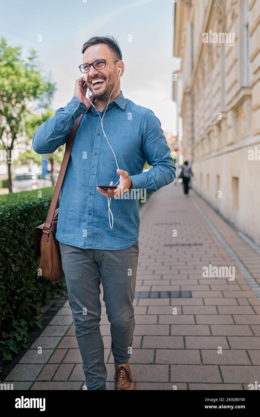 Homme adulte gai en chemise bleue, tenant son téléphone, tout en parlant et en écoutant à l'aide de ses écouteurs, à l'extérieur. Banque D'Images