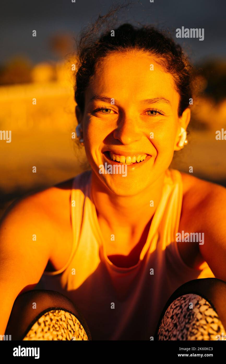Portrait d'une jeune femme souriante dans des vêtements de sport assis sur la plage et en écoutant de la musique sur des écouteurs avant de s'y rendre Banque D'Images