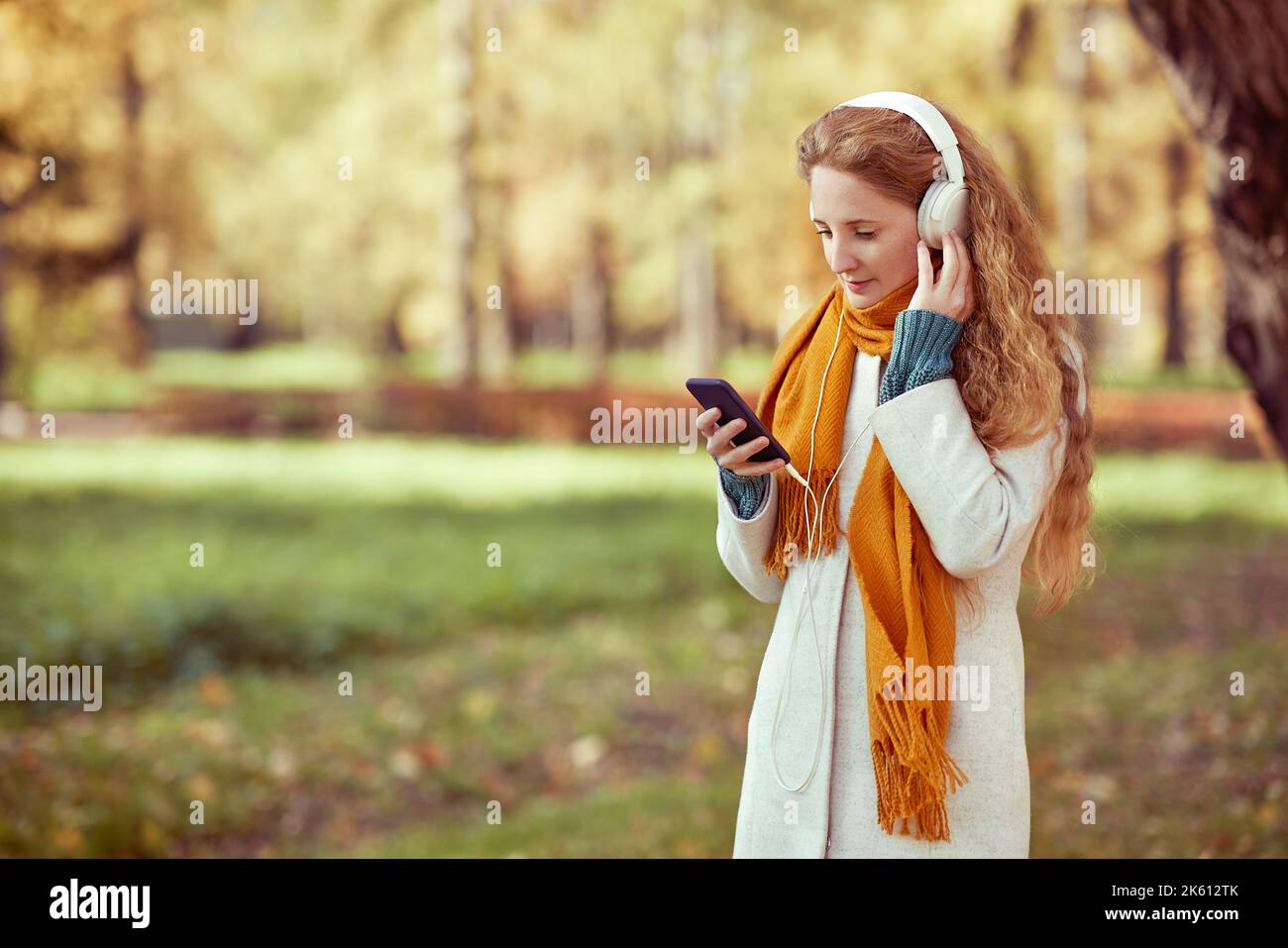 Une jeune femme aux cheveux rouges vêtue d'un manteau blanc marche dans un parc d'automne. Une fille écoute de la musique dans la forêt d'automne Banque D'Images