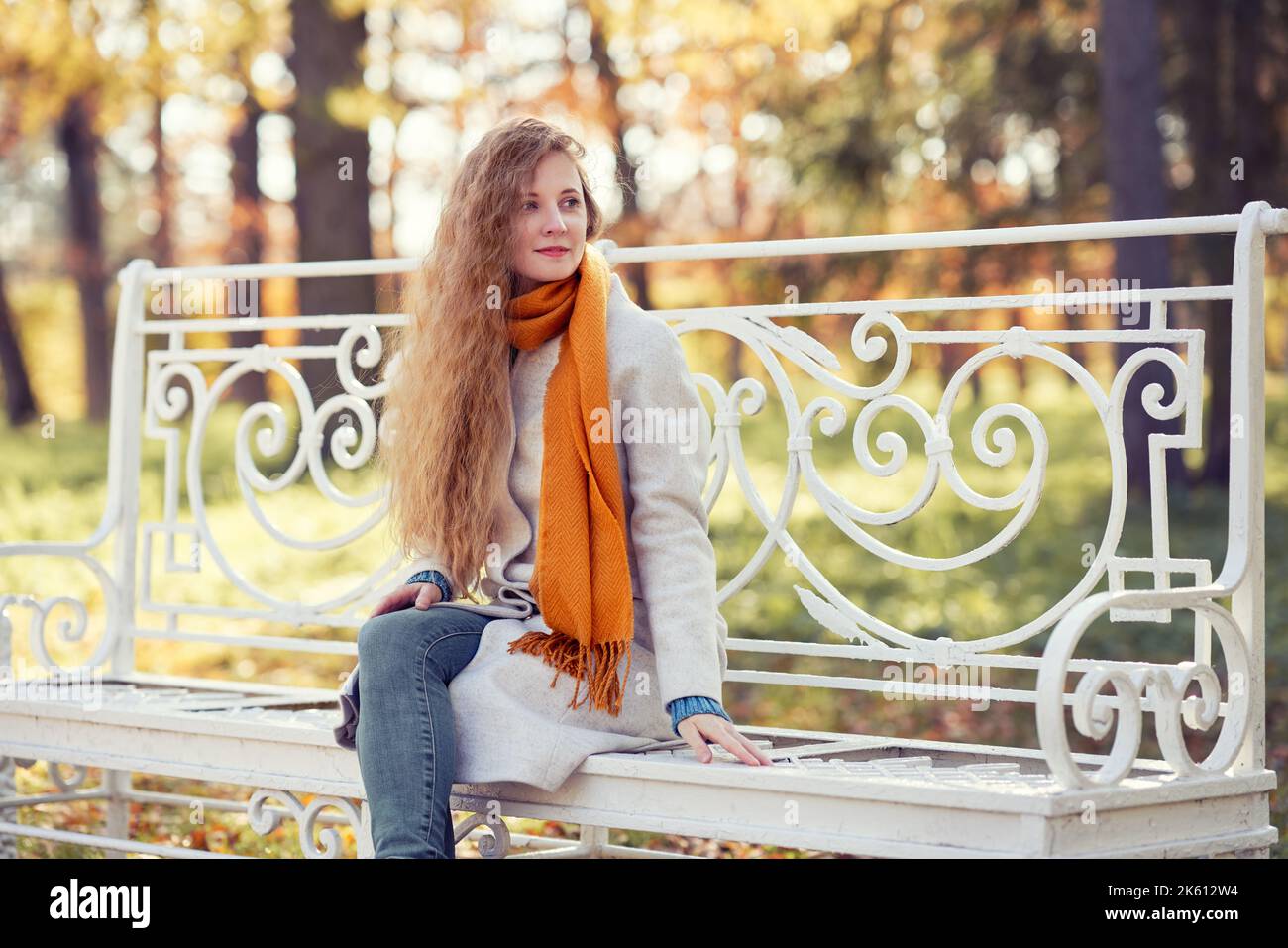 Une jeune femme aux cheveux rouges est assise sur un banc dans un parc d'automne. Une fille en manteau blanc se repose dans la forêt d'automne Banque D'Images