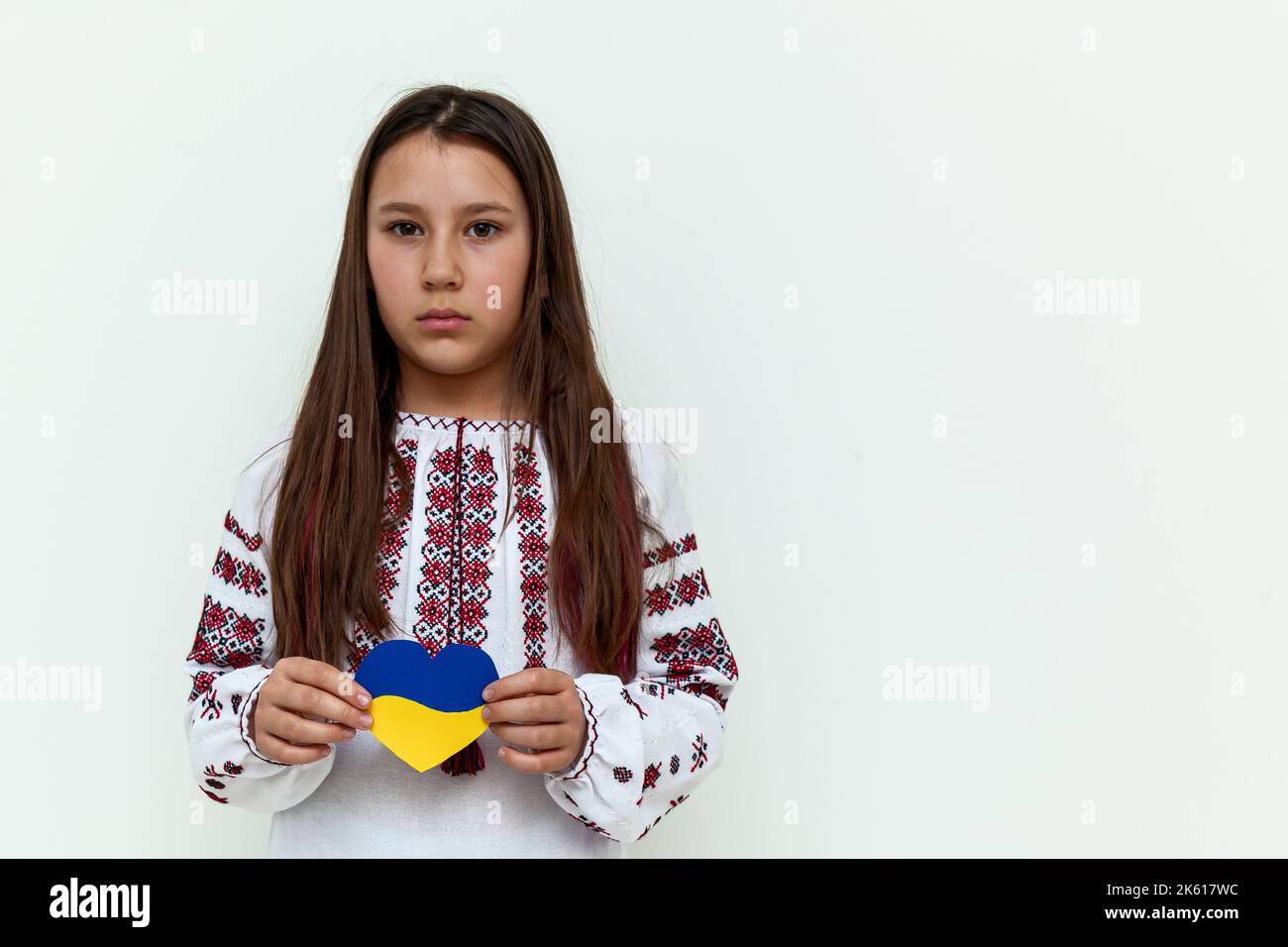 Une fille dans une robe brodée se tient contre un mur blanc, tenant un coeur peint en bleu et jaune. La jeune fille proteste contre la guerre et montre lo Banque D'Images