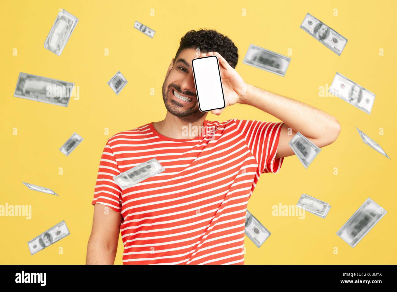 Investissement en ligne, le gagnant a surjoyé homme riche et gagner de l'argent. Un gars satisfait couvrant son oeil avec mobile vide exposition et se réjouissant de sa richesse. Studio d'intérieur isolé sur fond jaune. Banque D'Images