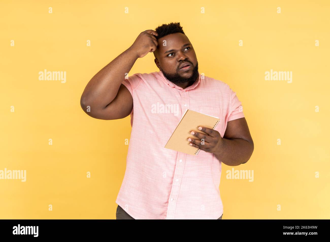 Portrait d'un homme attentionné portant une chemise rose pensant à une idée intelligente, tenant un crayon et un carnet pour écrire des plans, des listes de choses à faire. Studio d'intérieur isolé sur fond jaune. Banque D'Images