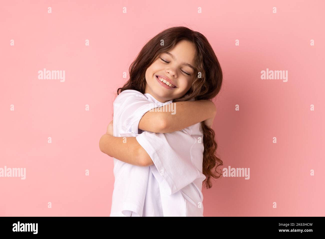 Portrait d'une petite fille heureuse satisfaite portant un T-shirt blanc s'embrassant, debout avec les yeux fermés du plaisir et du bonheur. Studio d'intérieur isolé sur fond rose. Banque D'Images