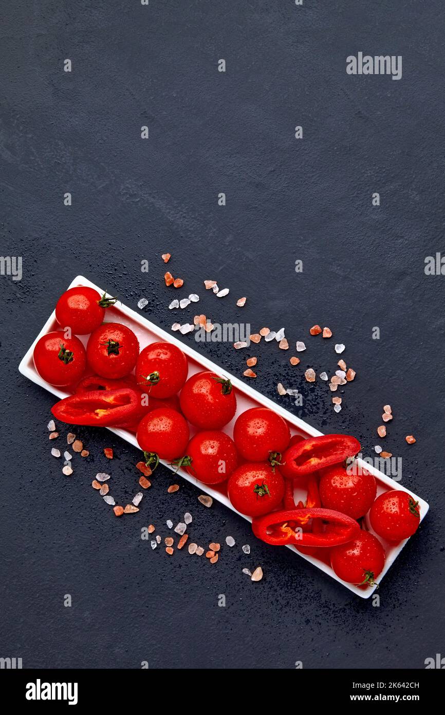 Tomates cerises fraîches lavées et tranches de paprika sur un plat oblong blanc avec du sel rose et des gouttes d'eau sur une table en béton noir Banque D'Images