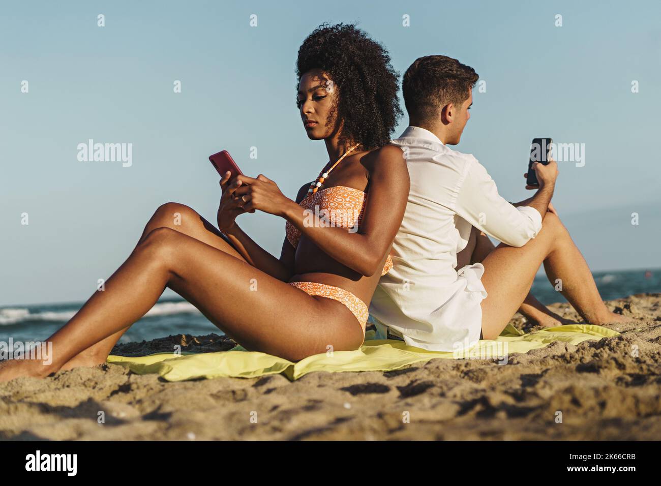Un jeune couple multiethnique qui se couche d'épaule à épaule sur la plage en été - les jeunes sont toujours connectés à Internet - la technologie Banque D'Images