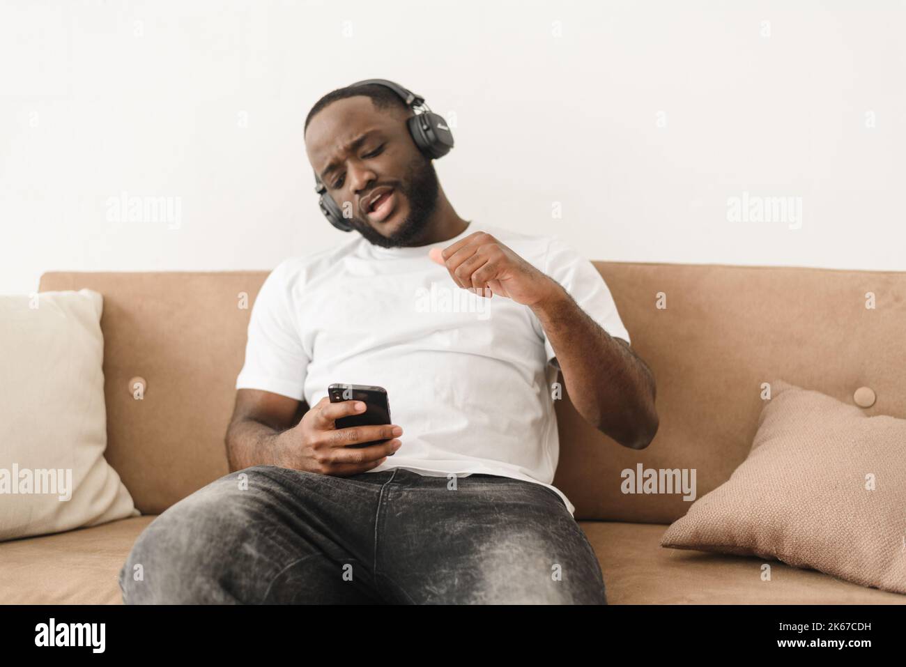 Jeune homme afro-américain qui écoute de la musique dans un casque sans fil tout en étant assis sur un canapé à la maison. Banque D'Images