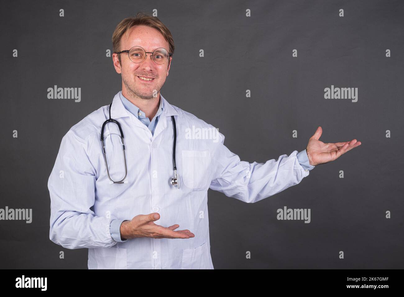 Homme heureux médecin avec stéthoscope sur le cou dans un pelage médical gestant sur le côté contre fond gris Banque D'Images