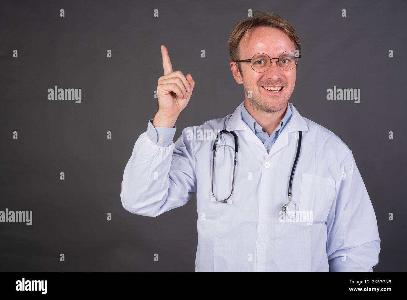 Homme heureux médecin avec stéthoscope sur le cou dans un pelage médical pointant le doigt sur fond gris Banque D'Images