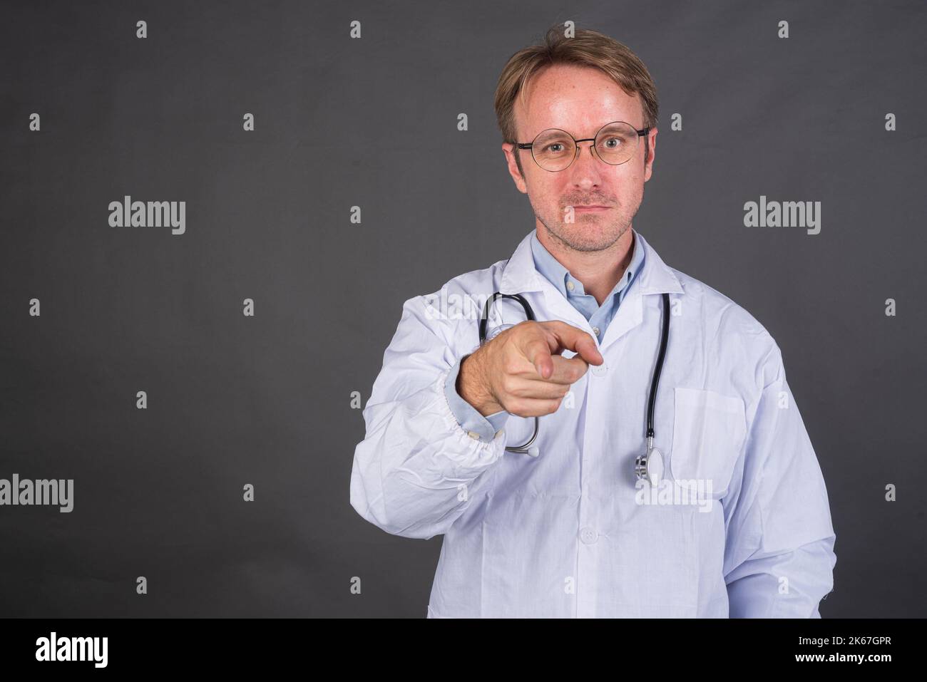Homme heureux médecin avec stéthoscope sur le cou dans un pelage médical pointant le doigt sur fond gris Banque D'Images