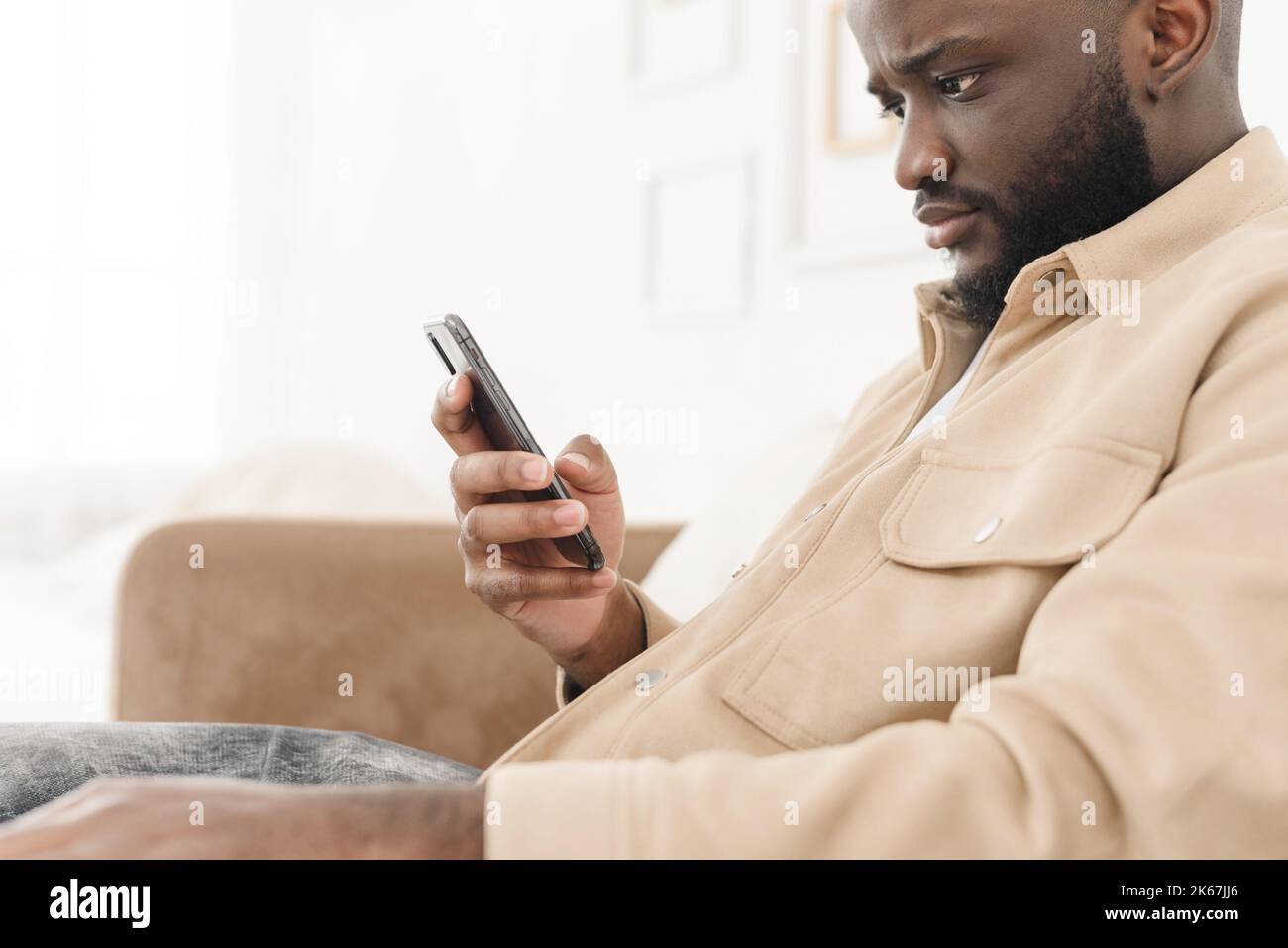 Un homme afro-américain agacé hurla au téléphone tout en étant assis sur le canapé à la maison. Un homme en colère regarde le téléphone tout en lisant de mauvaises nouvelles. Problèmes d'entreprise Banque D'Images