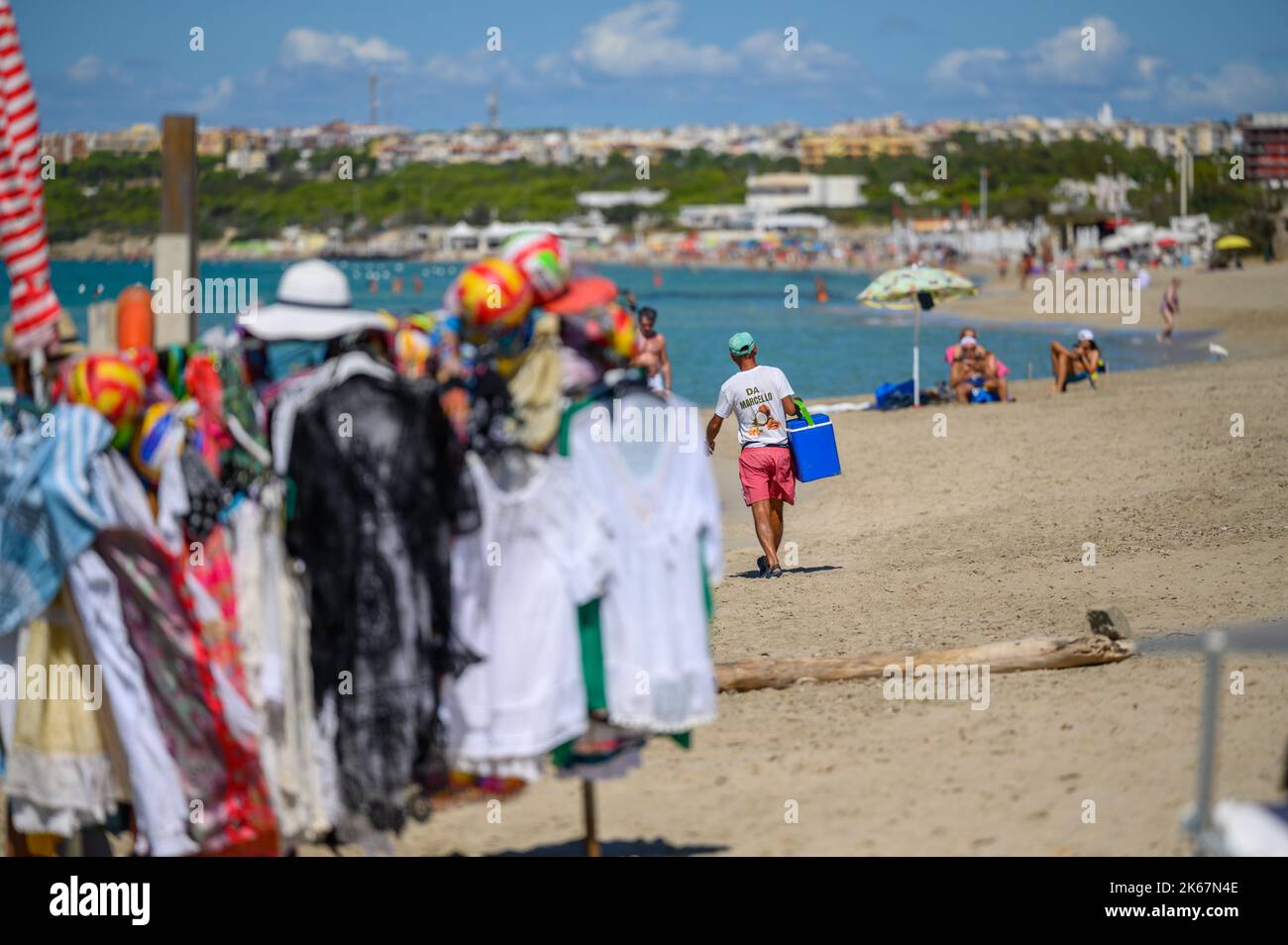 Un vendeur de plage marche avec des boissons froides à vendre avec plus de marchandises floues en premier plan sur la plage de Sottovento près de Gallipoli à Apulia (Puglia), Italie. Banque D'Images