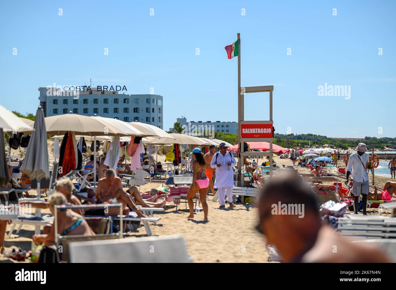 Un vendeur de plage se promène entre les baigneurs de soleil et les amateurs de plage sur la plage de Sottovento en essayant de vendre des colliers. Près de Gallipoli à Apulia (Puglia), Italie. Banque D'Images
