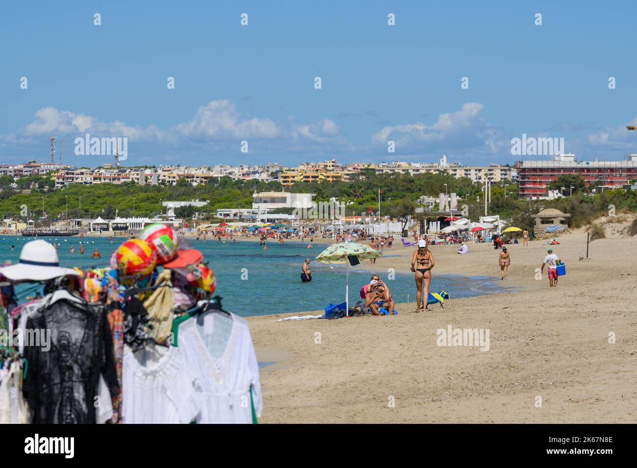 Vue de la plage de Sottovento vers Gallipoli avec les amateurs de plage, les baigneurs de soleil et les marchandises d'un vendeur de plage au premier plan. Pouilles (Puglia), Italie. Banque D'Images