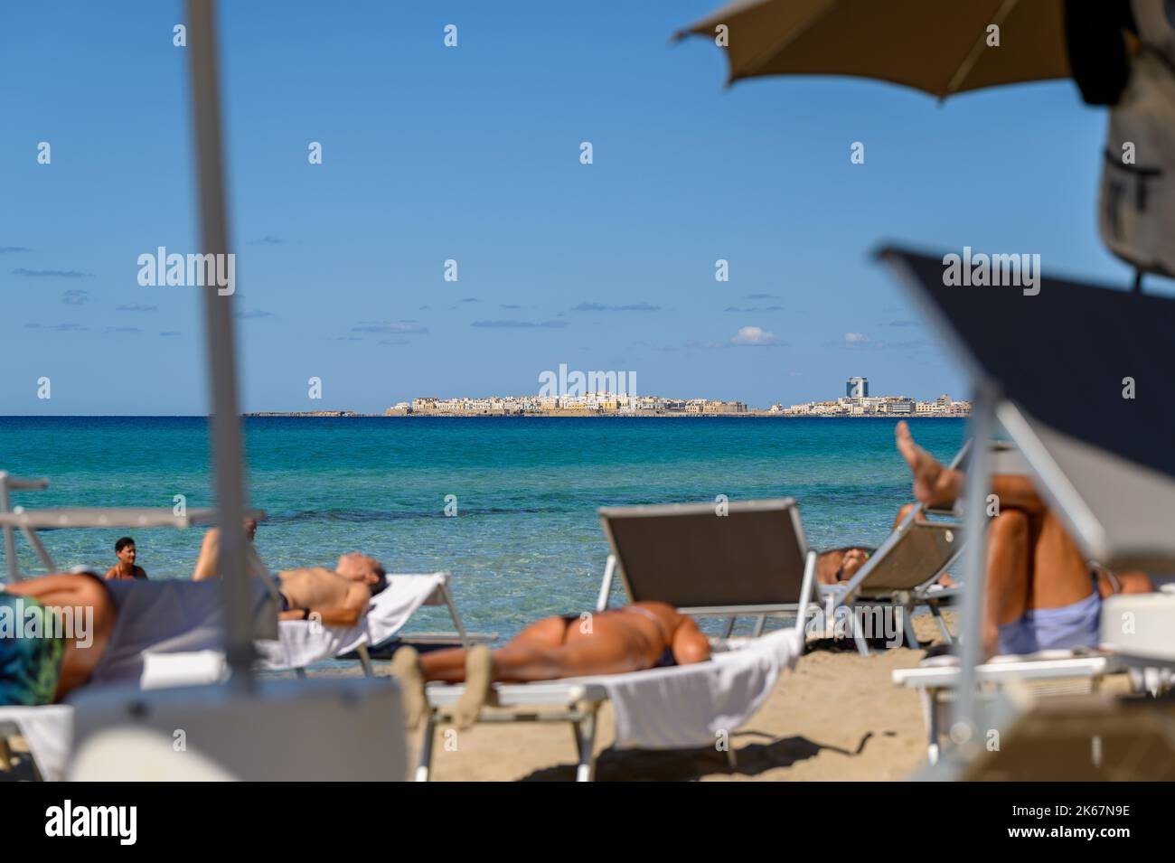 Ville de Gallipoli vue de l'autre côté de la baie depuis la plage de Sottovento avec des baigneurs de soleil hors de l'accent en premier plan. Pouilles (Puglia), Italie. Banque D'Images