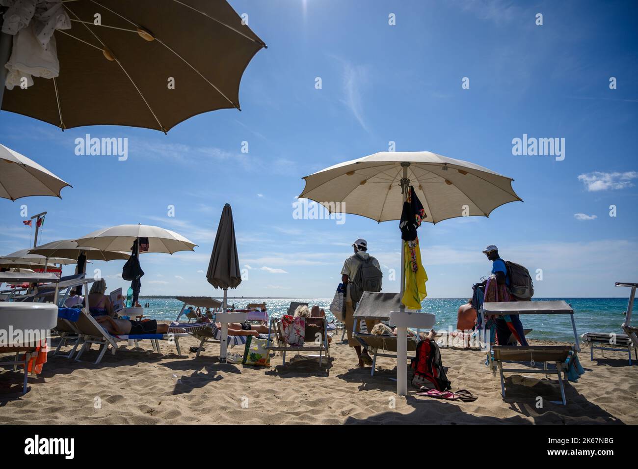 Deux vendeurs de plage qui marchent parmi les baigneurs offrant leurs produits à la plage de Sottovento vers la fin de la saison. Gallipoli, Pouilles, Italie. Banque D'Images
