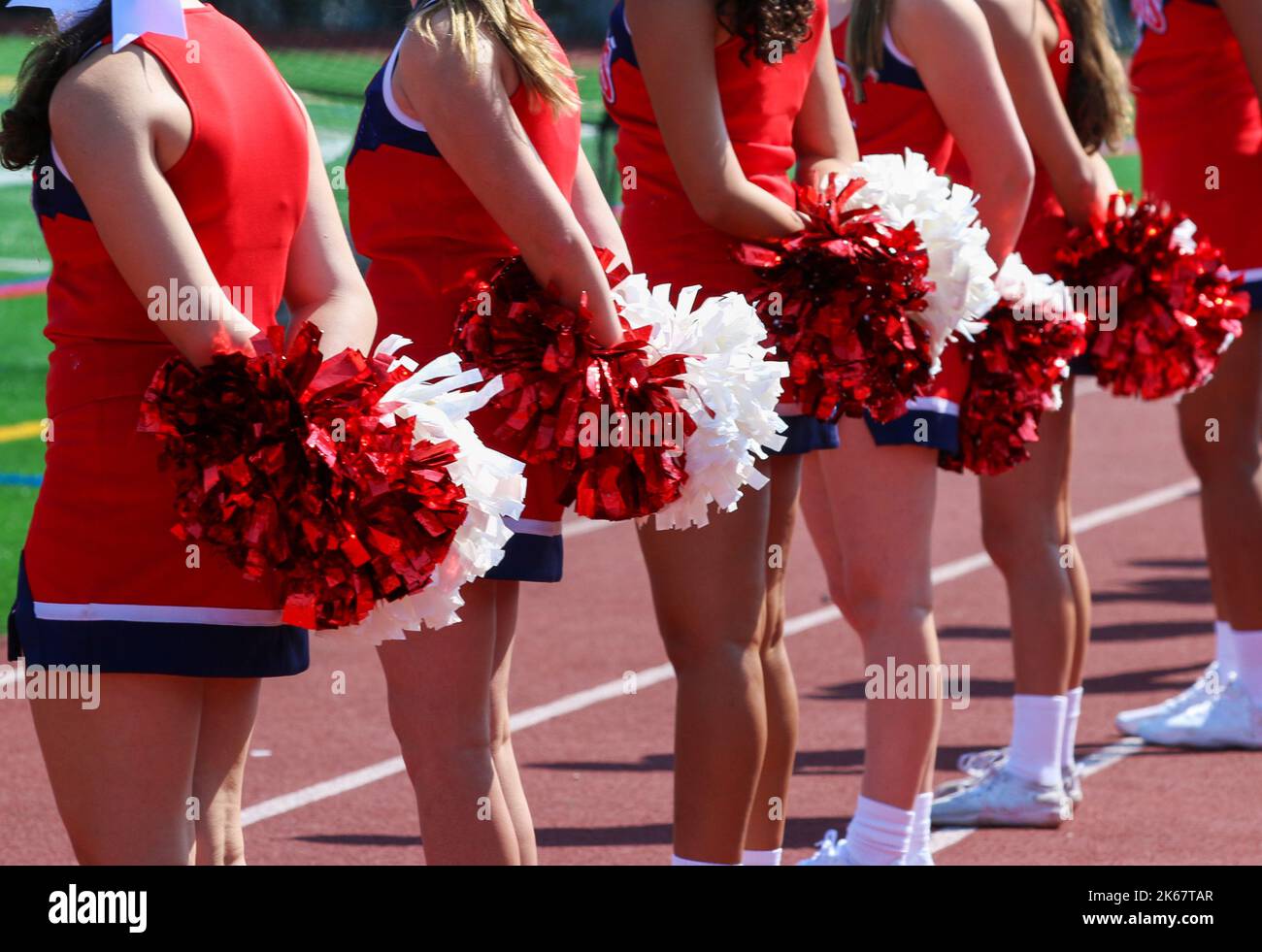 Les meneurs de l'école secondaire vêtus d'uniformes rouges debout sur le banc de touche tenant derrière eux leurs pompons rouges et blancs pendant un match de football. Banque D'Images
