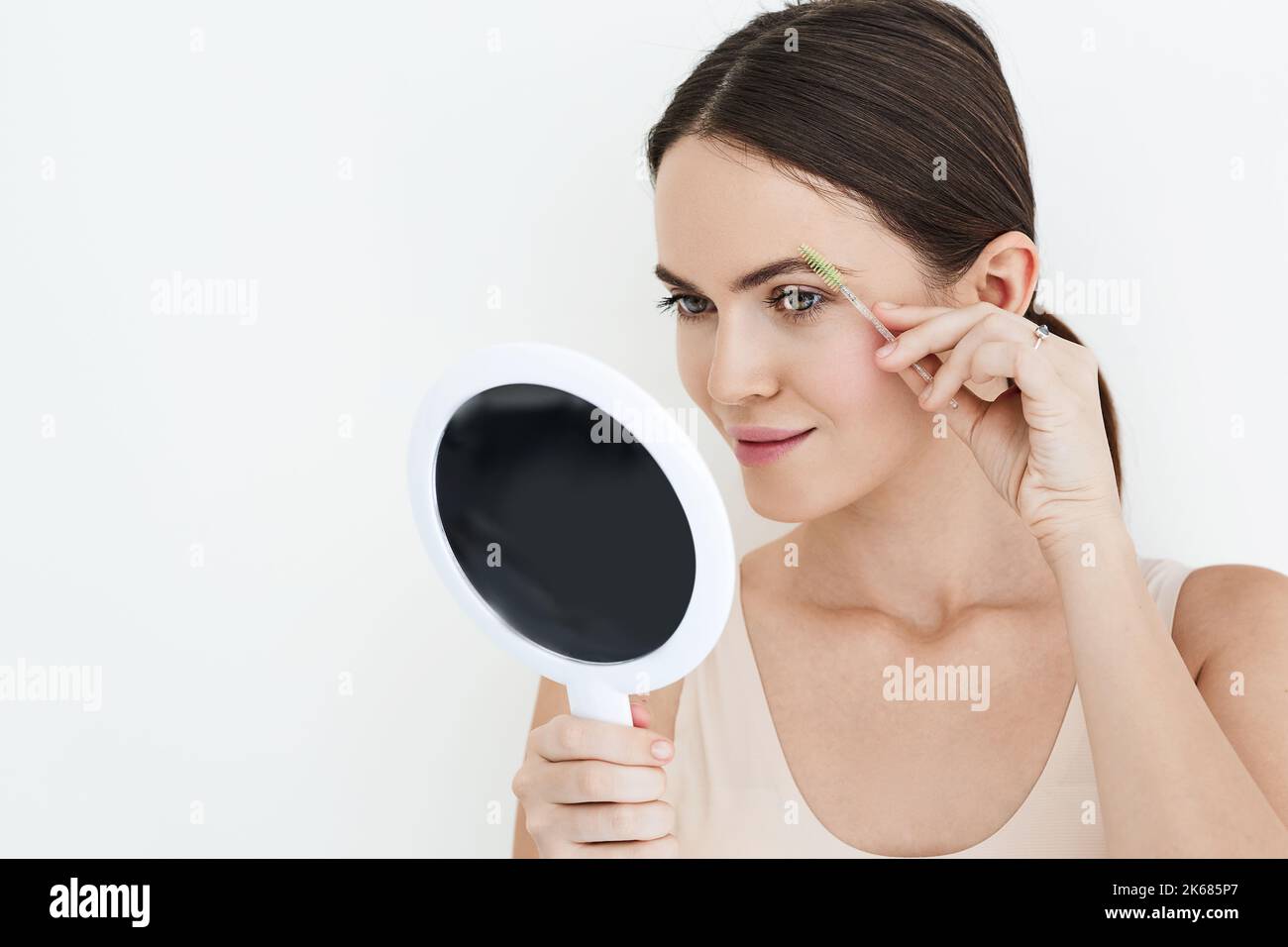 Belle femme peignant ses sourcils avec un outil de brosse tout en regardant dans le miroir portable, isolé sur blanc. Processus de brossage des sourcils Banque D'Images