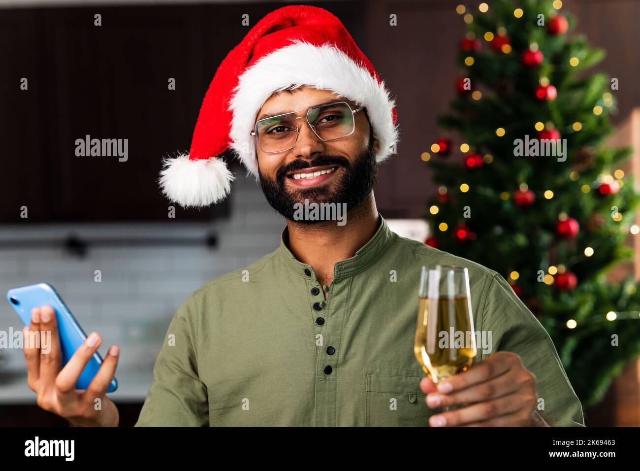 Homme indien boire de l'alcool à la maison arbre de Noël lumières guirlande arrière-plan et tenant smartphone Banque D'Images