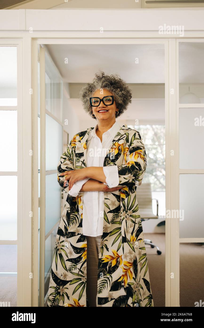 Femme d'affaires senior souriant à la caméra tout en se tenant dans un bureau avec ses bras croisés. Femme d'affaires heureuse travaillant dans un lieu de travail créatif. Banque D'Images