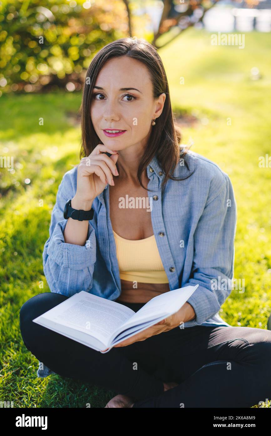 Belle femme sportive faisant des exercices de yoga et lire le livre assis sur l'herbe verte dans le parc du matin. Entraînement physique. Concept de passe-temps. Loisirs sportifs Banque D'Images