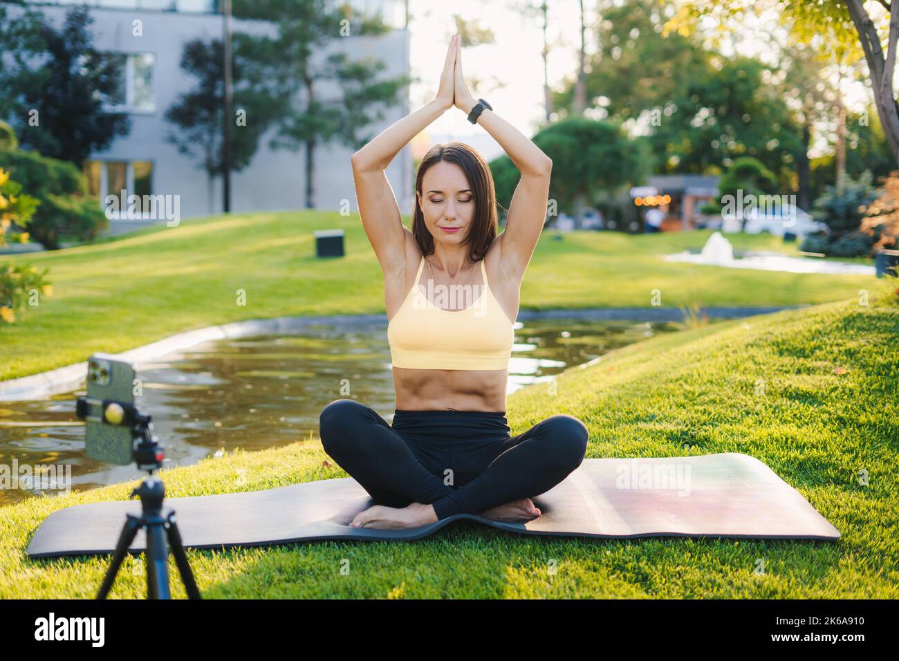 Portrait en gros plan de la jolie femme brune en vêtements de sport assise sur un tapis de yoga au parc et s'étirant tout en filmant sur un appareil photo extérieur Banque D'Images