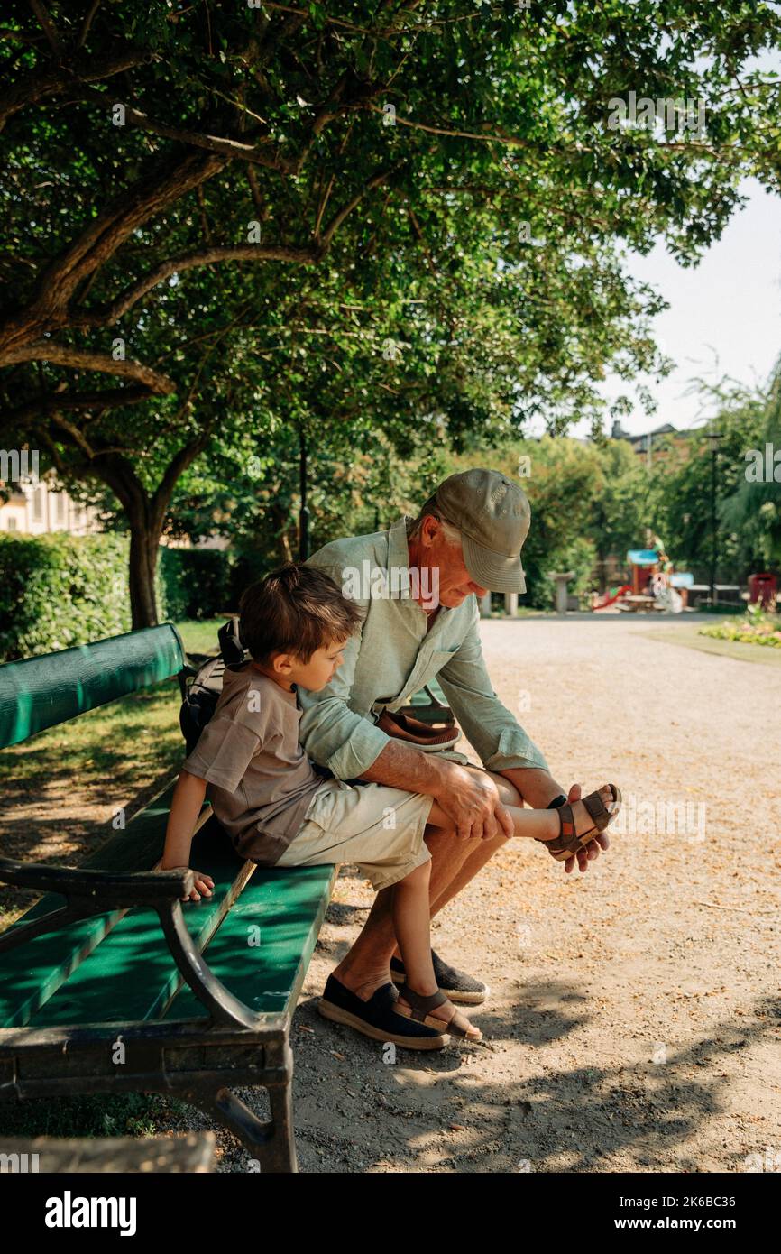Grand-père aidant le petit-fils à porter des sandales tout en étant assis sur un banc au parc Banque D'Images