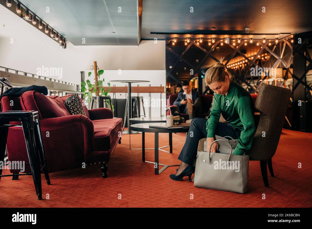 Femme d'affaires mûre cherchant dans le sac tout en étant assise sur une chaise au salon de l'hôtel Banque D'Images