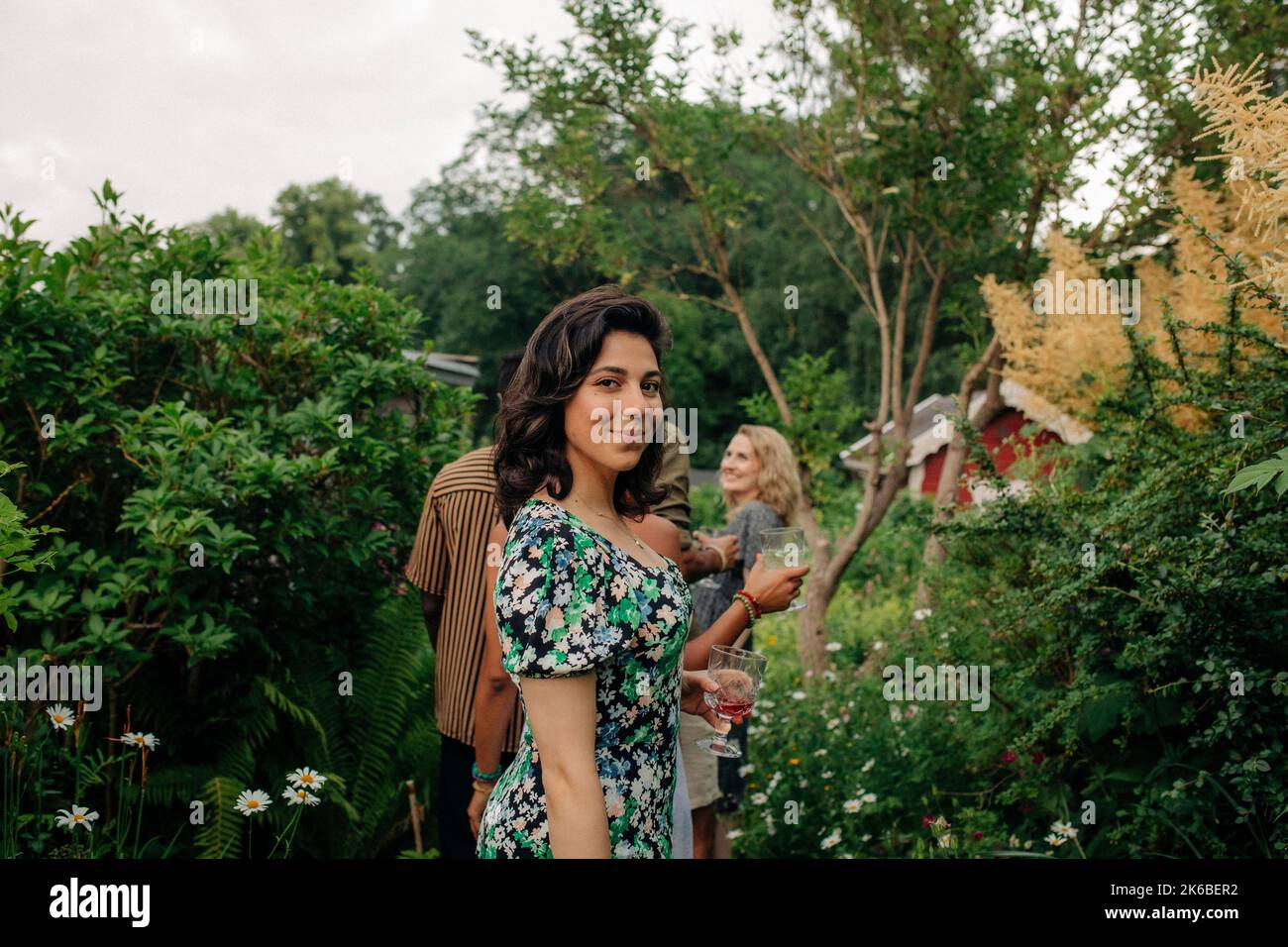Vue latérale d'une femme souriante avec boisson debout près des plantes dans le jardin Banque D'Images