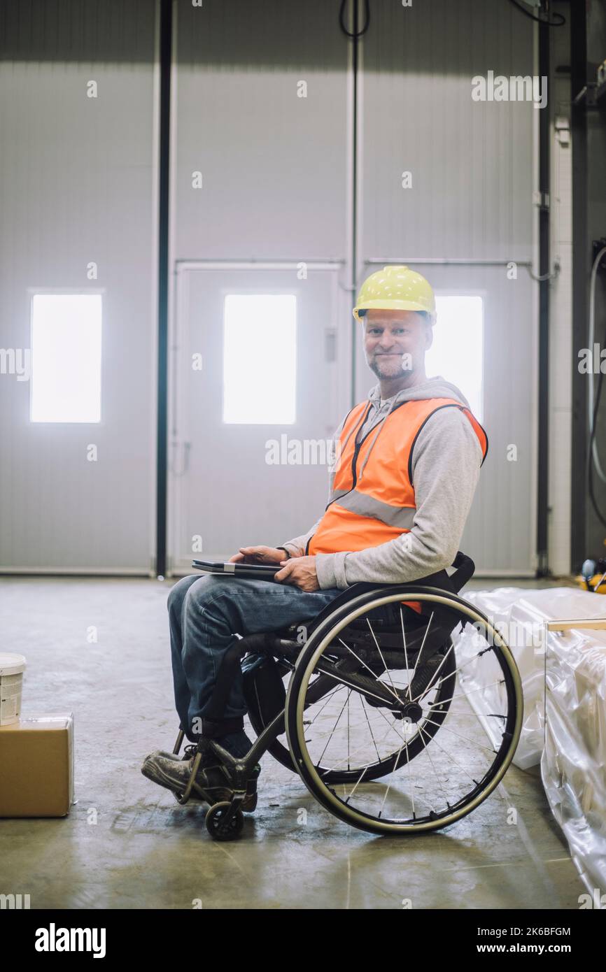 Portrait de menuisier souriant avec handicap tenant une tablette numérique tout en étant assis sur un fauteuil roulant dans un entrepôt Banque D'Images