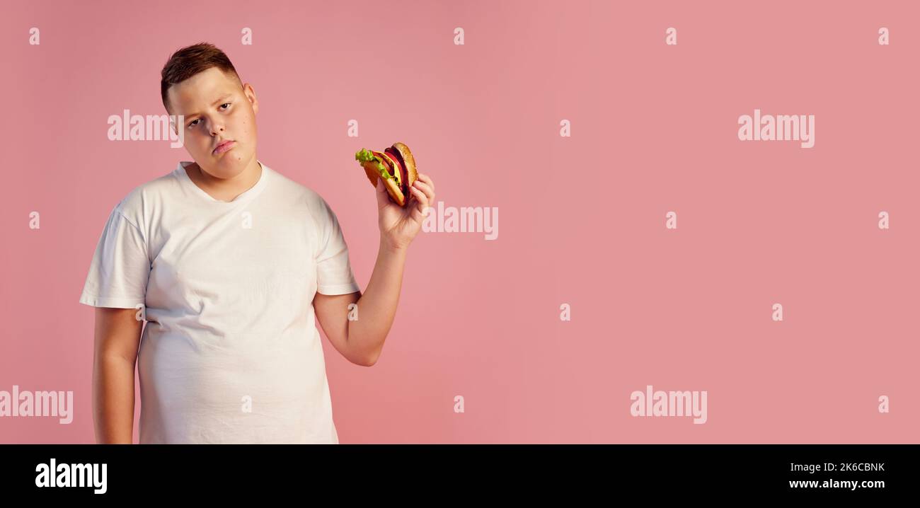 Fatigué agacé de poids excessif adolescent garçon dans le t-shirt blanc refuse de manger le gros hamburger isolé sur fond rose. Restauration rapide, goût, corps positif, émotions Banque D'Images