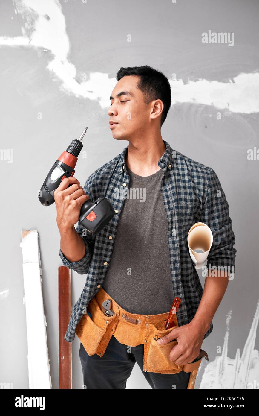 Un homme asiatique séduisant souffle une perceuse debout avec des outils de bricolage et une ceinture à outils Banque D'Images