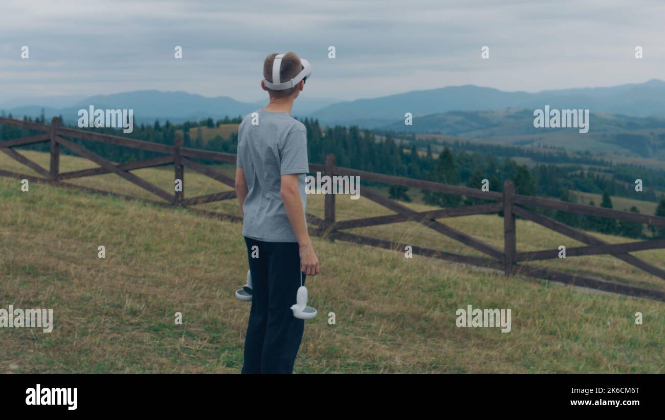 Jeune homme jouant à des jeux de réalité augmentée sur la nature, restant sur de belles collines verdoyantes. Les joueurs testant les lunettes VR et les contrôleurs sans fil, en passant du temps libre à l'extérieur Banque D'Images