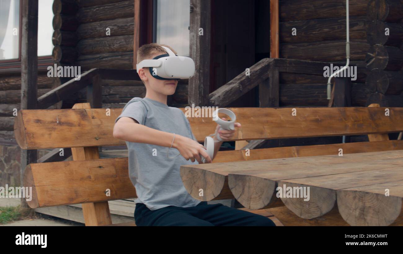 Jeune homme jouant à des jeux de réalité augmentée sur la nature, assis sur un banc en bois. Les joueurs testant les lunettes VR et les contrôleurs sans fil, en passant du temps libre à l'extérieur Banque D'Images