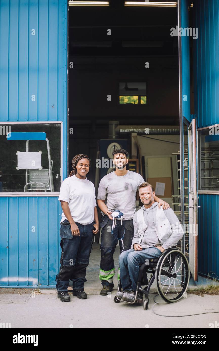 Pleine longueur de peintres hommes et femmes souriants avec un collègue assis en fauteuil roulant à l'extérieur de l'atelier Banque D'Images