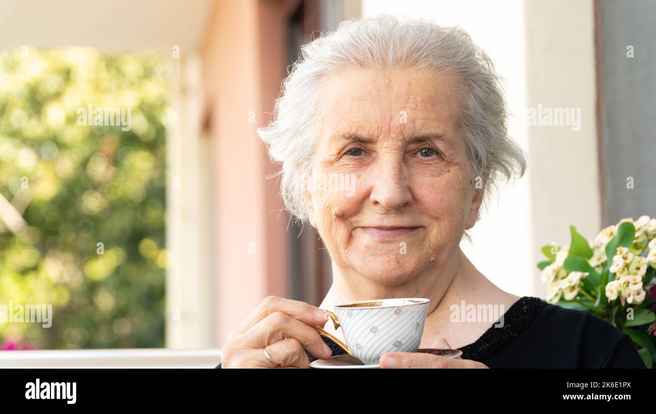 Vieille femme souriante regardant un appareil photo, buvant un café turc et se détendant sur le balcon. Femme âgée insouciante en 70s souriant et appréciant le temps. Banque D'Images
