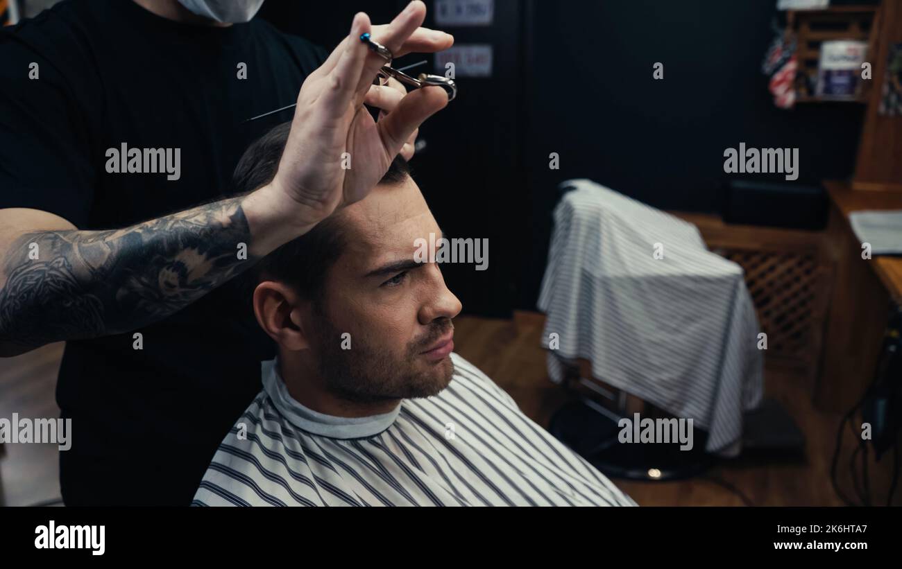 Coiffeur tatoué avec des ciseaux coupant les cheveux de l'homme dans le cap de coiffure en salon, image de stock Banque D'Images