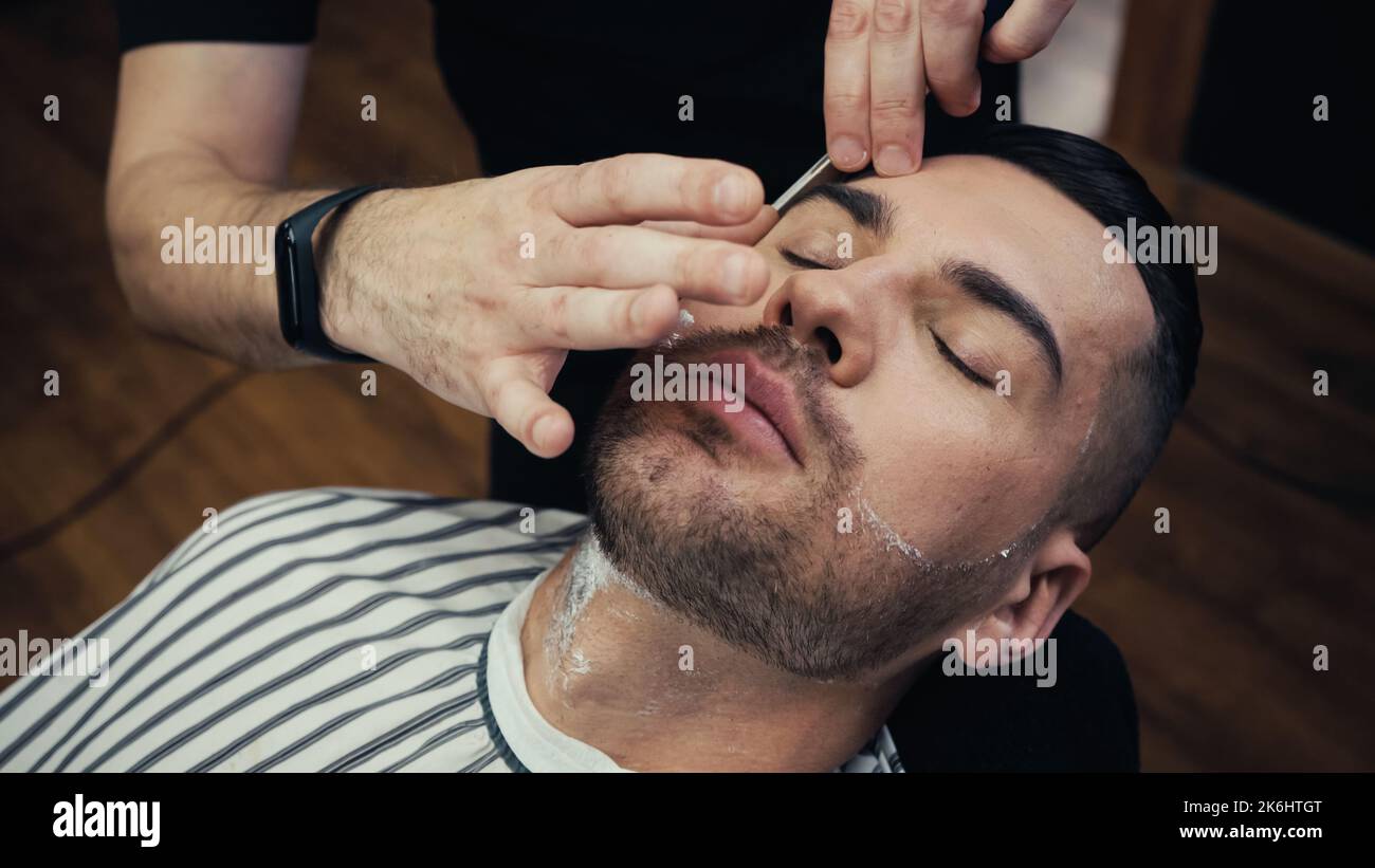 Vue en grand angle de la barbe de rasage de coiffeur du client dans le cap de coiffure dans le barbershop, image de stock Banque D'Images