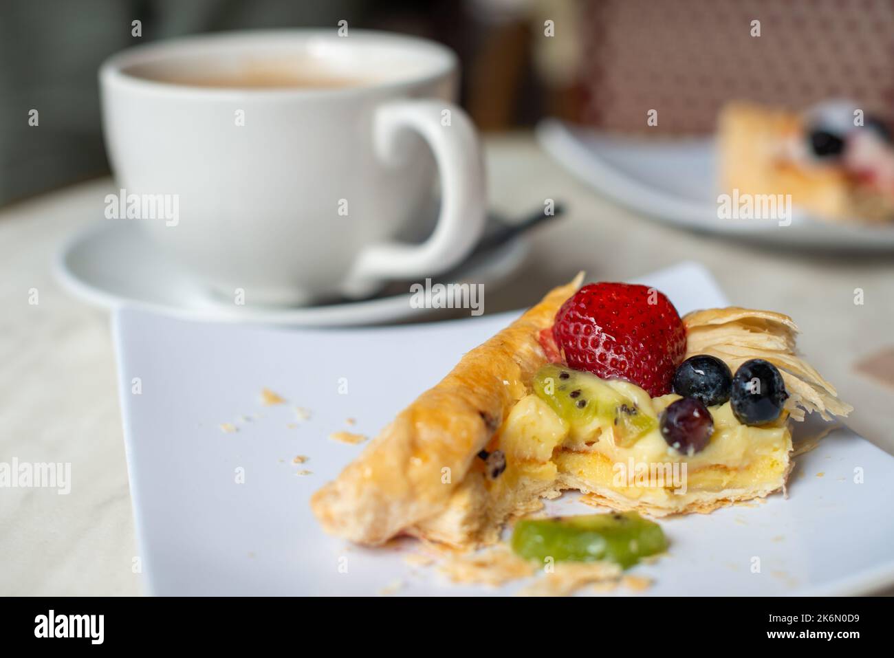 Une tasse en porcelaine blanche et une soucoupe avec une petite cuillère à café sur la soucoupe. La tasse est remplie de café chaud et de crème. La plaque a une pâtisserie avec des baies. Banque D'Images