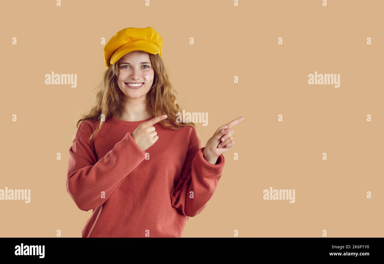 Jeune femme souriante portant un sweat-shirt rouge et un chapeau pointant sur l'espace de copie sur fond beige. Banque D'Images